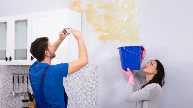 Homeowner photographing water damage on a wall while another holds a bucket